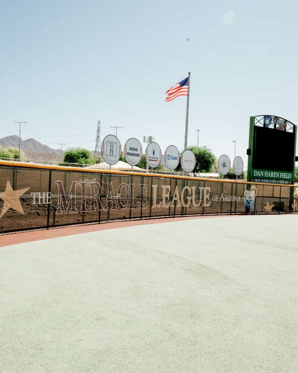 Close-up of the Miracle League of Arizona signage on the outfield fence at Dan Haren Field, with the American flag flying and players on the field. Featured in Scottsdale Lifestyle Magazine.